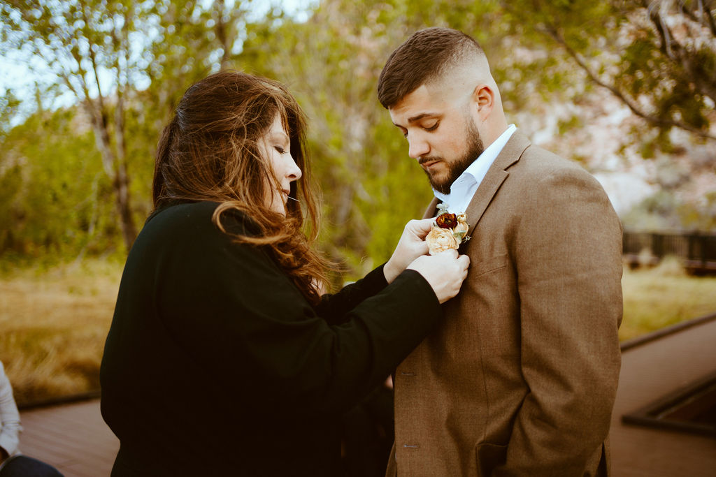 Rustic Boho Never Looked So Good! The mother of the groom helps him place a toffee and deep brown boho styled boutonniere on his lapel. Her hair bowls in the wind. His hair has a fresh fade going into a neatly trimmed beard. 