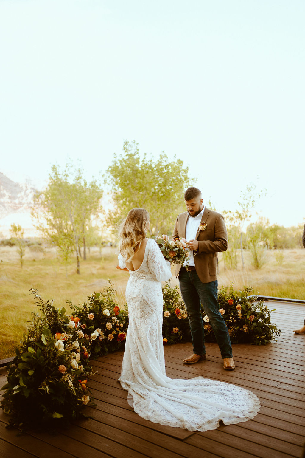 Seeing the grooms front as he reads the vows. He wears a white shirt with no tie and brown suit jacket. Dark fitted blue jeans and glossy tan dress shoes. The bride faces him. The back white lace wedding dress with puffy sleeves swoops low down her back. 