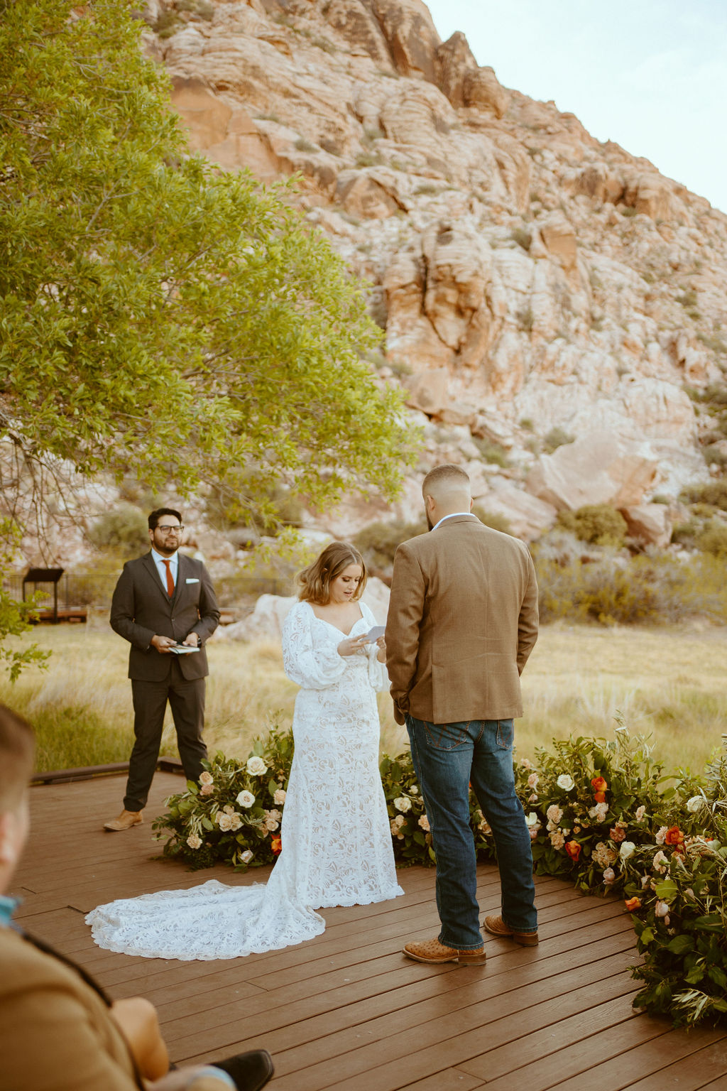 The Bride reads her vows to the groom. Her fitted white lace dress and small train cascades around her. The rustic floral ground arch encircles the bride and groom. The groom is facing the bride in his brown suit jacket with dark blue jeans. 