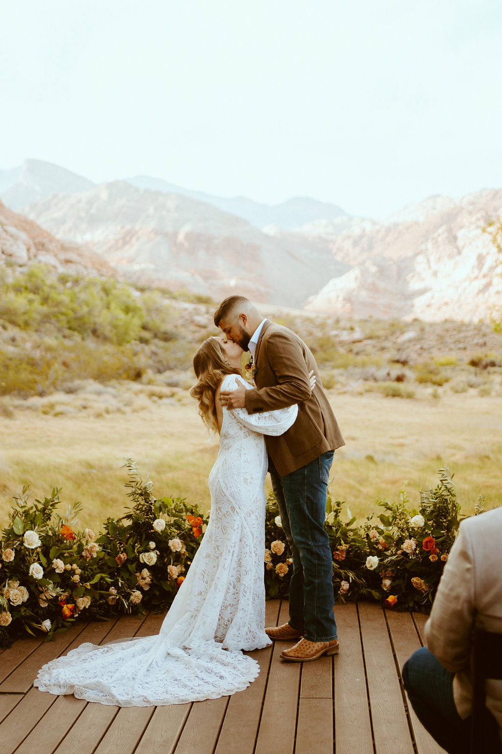 The bride and groom share their first kiss as husband and wife. The desert mountains are in the background. The groom holds the brides shoudlers of her whtie fitted lace with puffy sleeves wedding dress. 