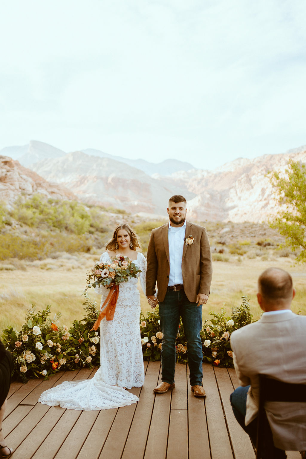 Rustic Boho Never Looked So Good! Hand in hand the bride and groom smile at their friend and family. The bride holds her rustic boho bridal bouquet with burnt orange lace wrapped around the stems. Their rustic floral ground arch behind them. 