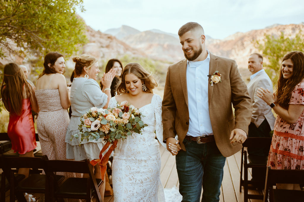 Rustic Boho Never Looked So Good! The wedding party cheers and the bride and groom walk down the isle hand in hand. You can see close up the white lace details of the brides dress and her bridal bouquet with dahlias, roses, and eucalyptus The bride and groom smile happily. 