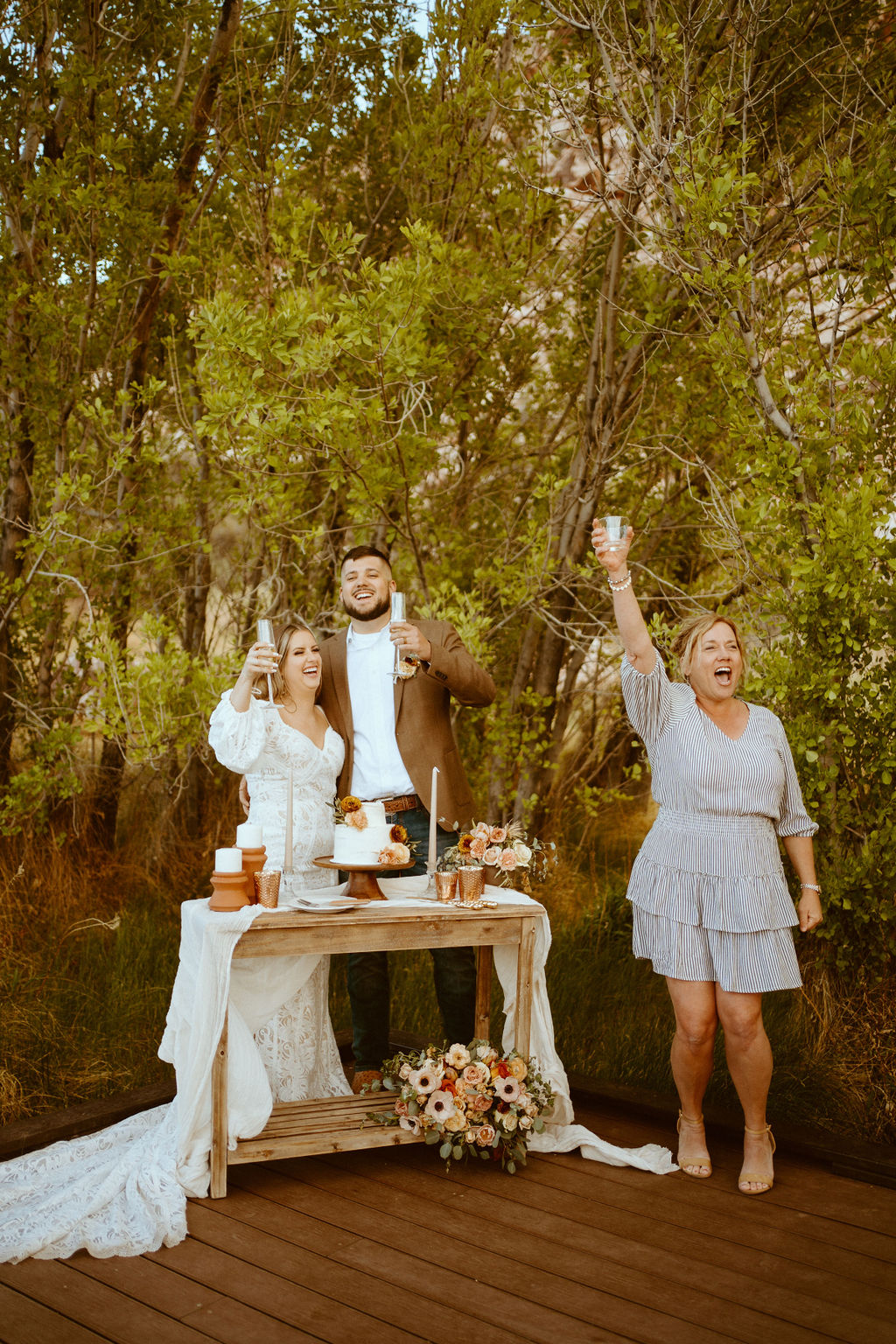 A wedding party guest standing next to the bride and groom at their cake table cheering excitedly with her glass way up in the air. The bride and groom laughing as they raise their glasses to cheer. 