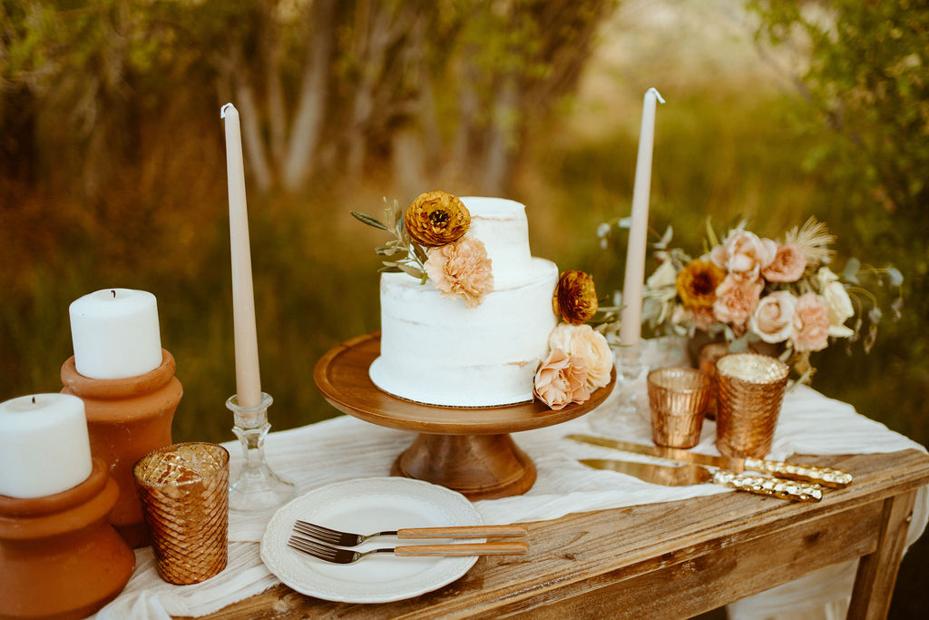 Rustic Boho Never Looked So Good! Close up of the cake table. With Ceramic and crystal candle holders. A rustic boho floral display and golden cake cutter. The white simple two tiers cake with boho florals decorated on top. 