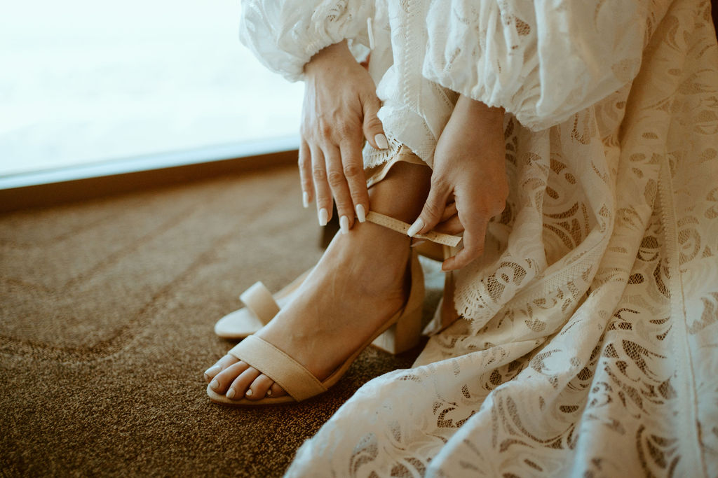Close up of the bride clipping on her short heeled beige shoes. Her white manicured nails push the heel strap together. Her lace dress and sleeves just barely seen in the close up. 