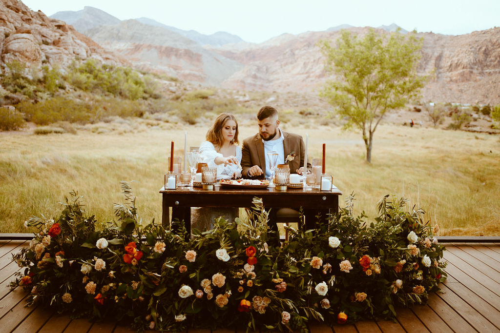 Rustic Boho Never Looked So Good! Couple sit at their sweetheart table to enjoy their charcuterie board together. Their ground rustic boho floral arch wraps around the dark wooden table. 