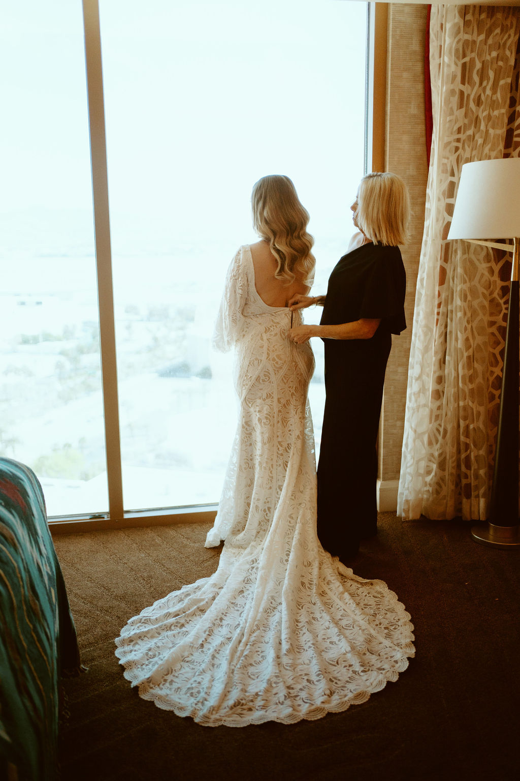 Bride and her mother stand at the hotel windows. The mother of the bride helps zip up her dress as she gets ready to head to her elopement. The brides lace train cascades behind her. 