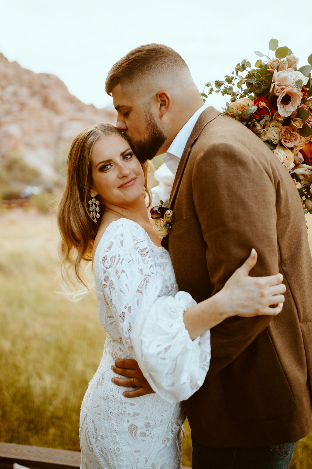 Rustic Boho Never Looked So Good! The groom in his dark brown suit jacket kisses the brides temple. The bride looks at the camera in a soft smile as she holds her bridal bouquet. She wears a white fitted lace dress with long puffy sleaves. 
