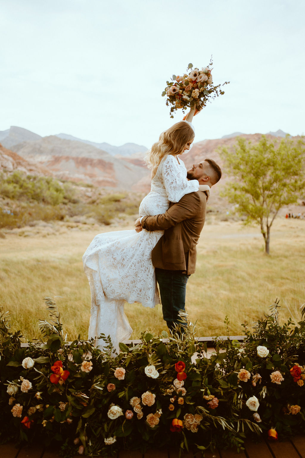 Groom lifts bride up in the air as she holds her bridal bouquet over her head. Their rustic boho ground floral arch wraps around them as the brides white lace dress hangs down. 