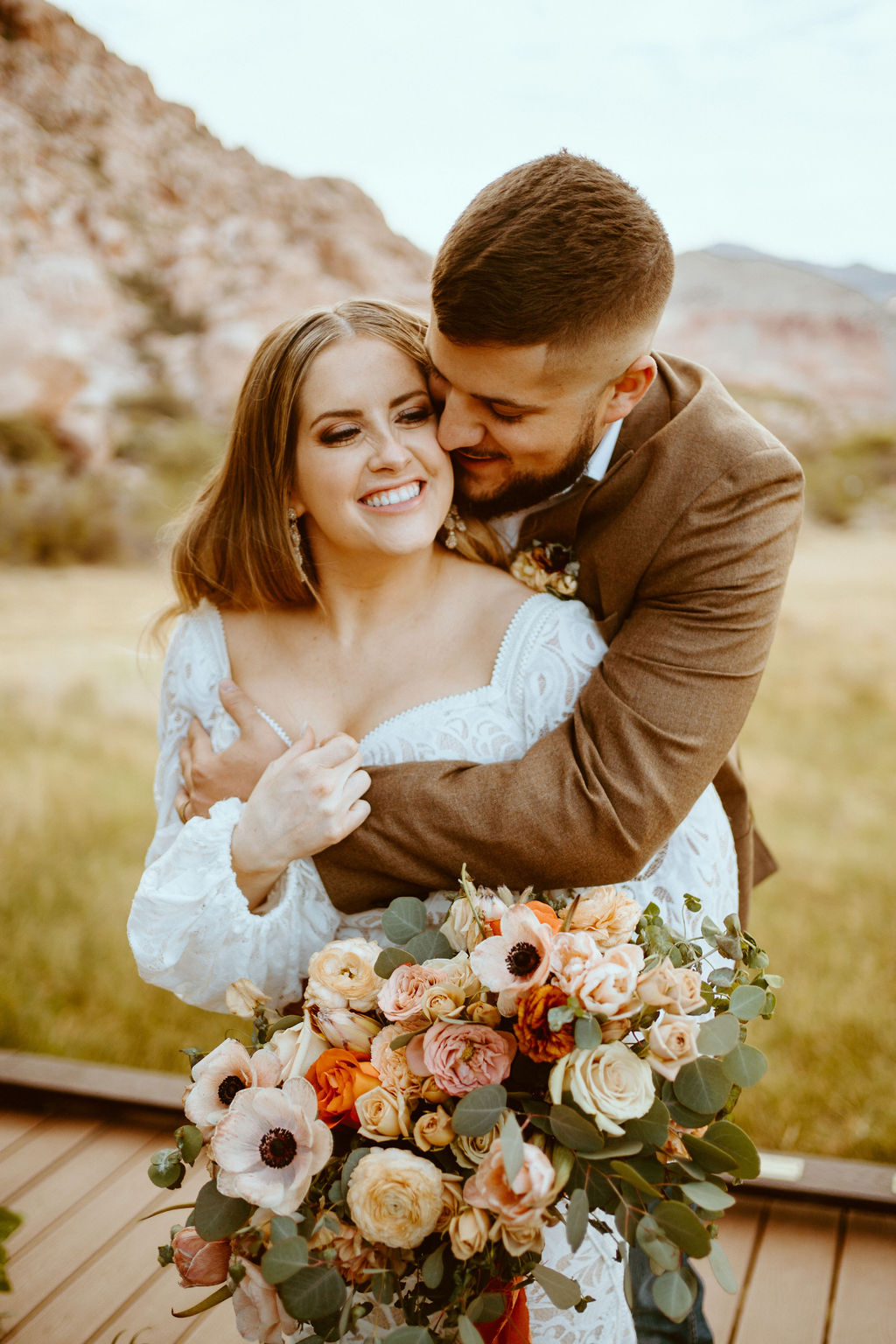 Rustic Boho Never Looked So Good! Groom has his arms wrapped around the bride holding her close nuzzling her cheek. She smiles as she holds her rustic boho bridal bouquet. 