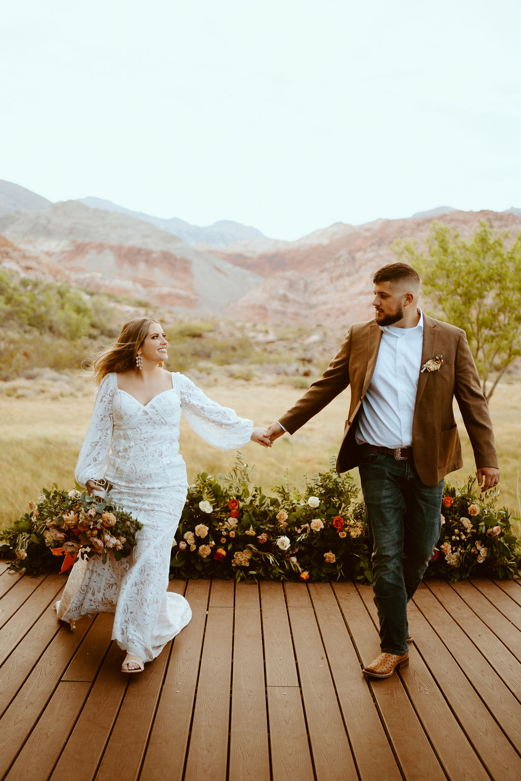Rustic Boho Never Looked So Good! Holding hands and walking across a wooden boardwalk. Rustic boho floral ground arch is behind the couple as she holds her rustic bridal bouquet. 