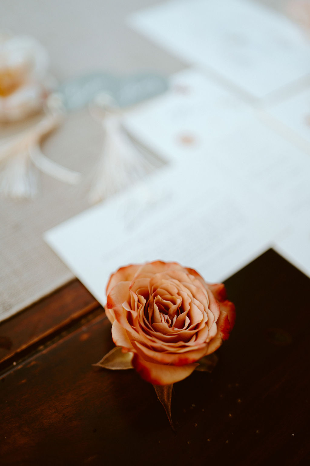Close up of a burnt orange, red, and pink rose placed along with the wedding stationary blurry in the background. 