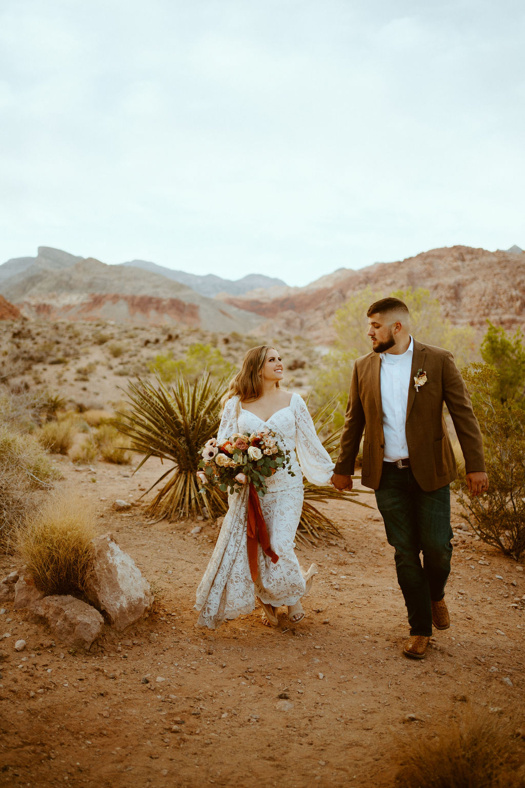 Rustic Boho Never Looked So Good! Walking hand in hand in the Las Vegas Desert with the Red Rock mountains behind. The bride wears a fitted white lace boho styled dress with large puffy sleaves. She holds a rustic orange, peach, pink and toffee bridal bouquet. The groom wears a dark brown suit jacket with dark fitted blue jeans and white shirt. 