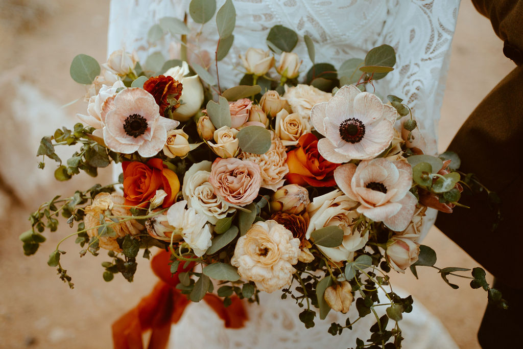 Rustic Boho Never Looked So Good! Close up of the brides wedding bouquet. You can see the rich colors of bright burnt orange roses, with toffee, peach, pink, and brown roses. Soft white with a little pink dahlias sprinkled through the bouquet. Eucalyptus leaves fill in the greenery of the bouquet. 