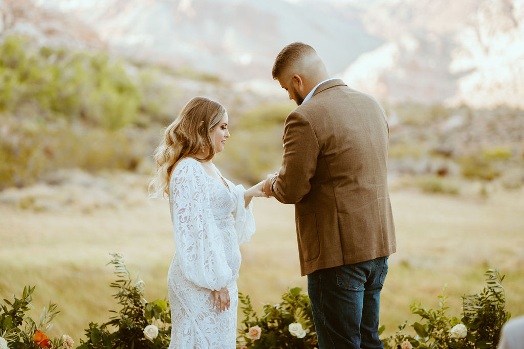 Rustic Boho Never Looked So Good! The groom in his dark brown suit jacket places a ring on his brides finger. Her fitted white lace and puffy sleeves wedding dress stands out against the green and yellow background. 