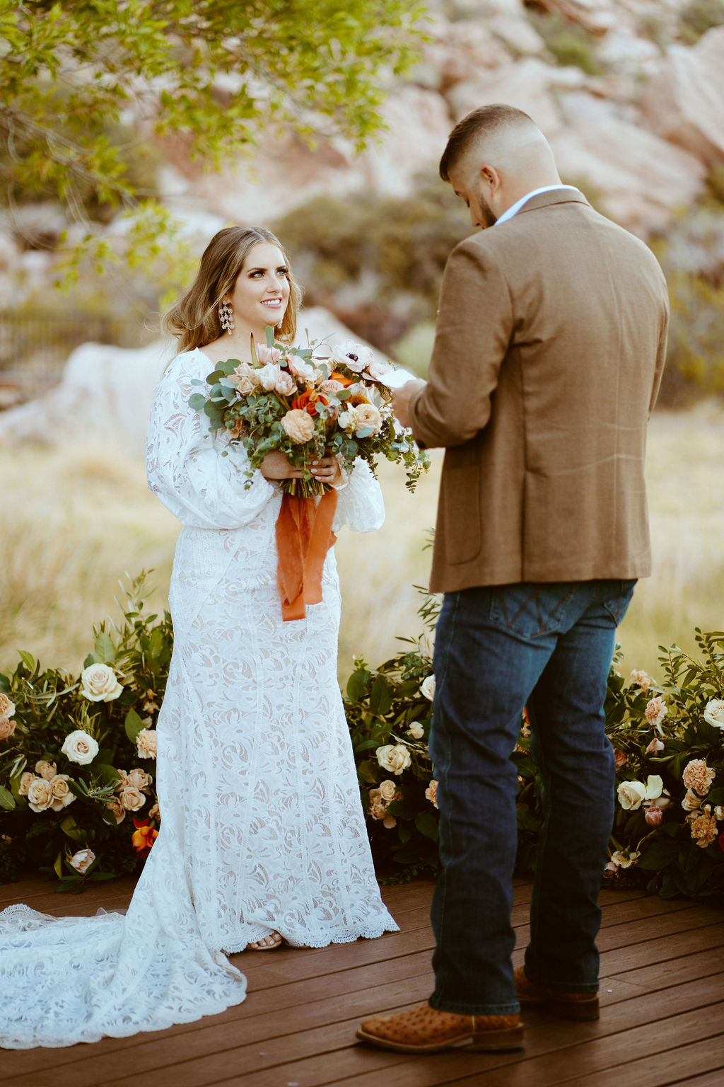 Rustic Boho Never Looked So Good! Close up of the bride with the grooms back facing the camera. She holds her rustic boho bridal bouquet with long burnt orange ribbon flowing down the bouquet. Wearing fitted white lace wedding dress and puffy sleeves. The groom reads his wedding vows. 