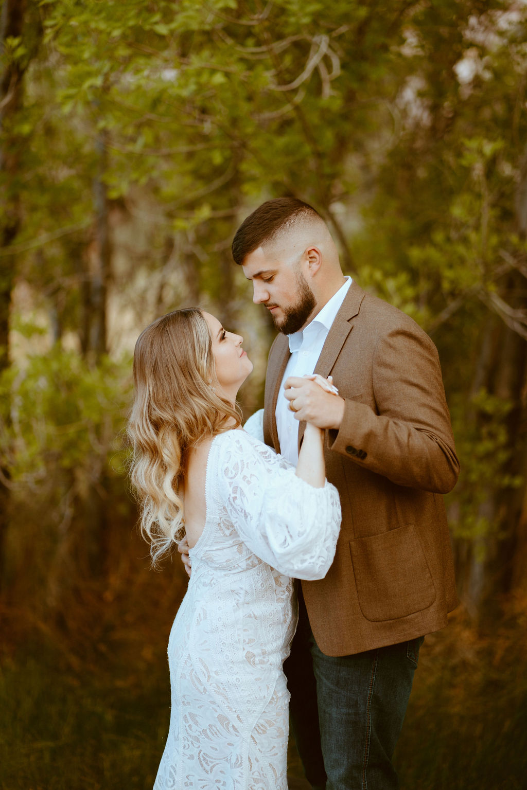 Rustic Boho Never Looked So Good! Bride and groom hold hands as they share their first dance. Bride is wearing a fitted white lace wedding dress with puffy sleeves. Groom is wearing a dark brown suit jacket, white shirt with no tie, and fitted dark blue jeans. 