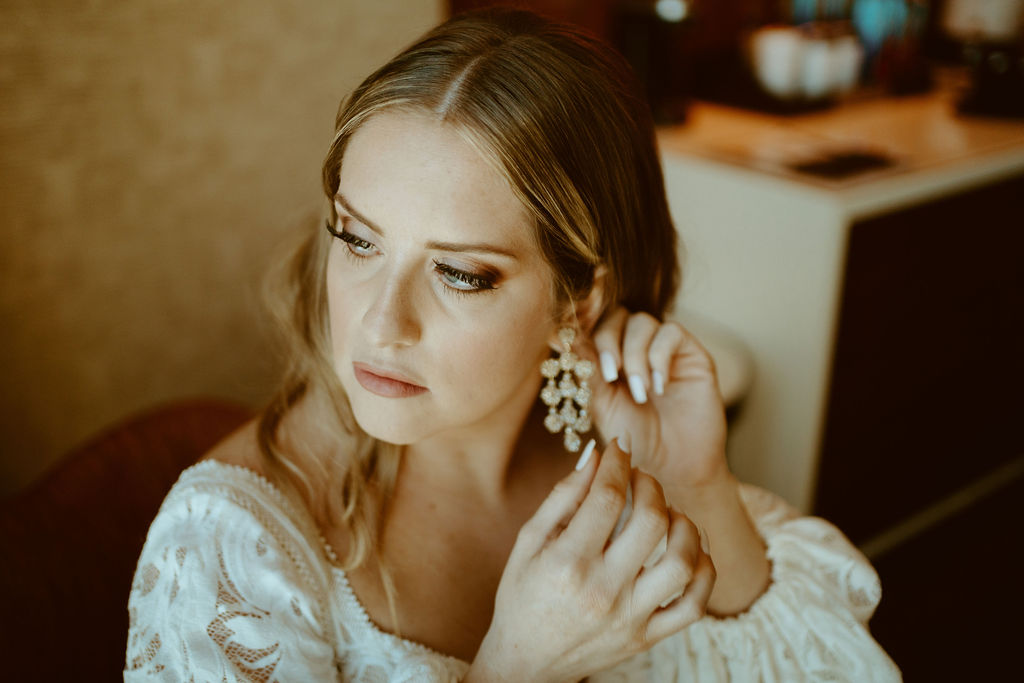 Rustic Boho Never Looked So Good! The bride looks off to the side as she places a long dangling crystal earing in. She wears a soft smokey eye make up and her hair is pulled away from her ear in long Hollywood curls.
