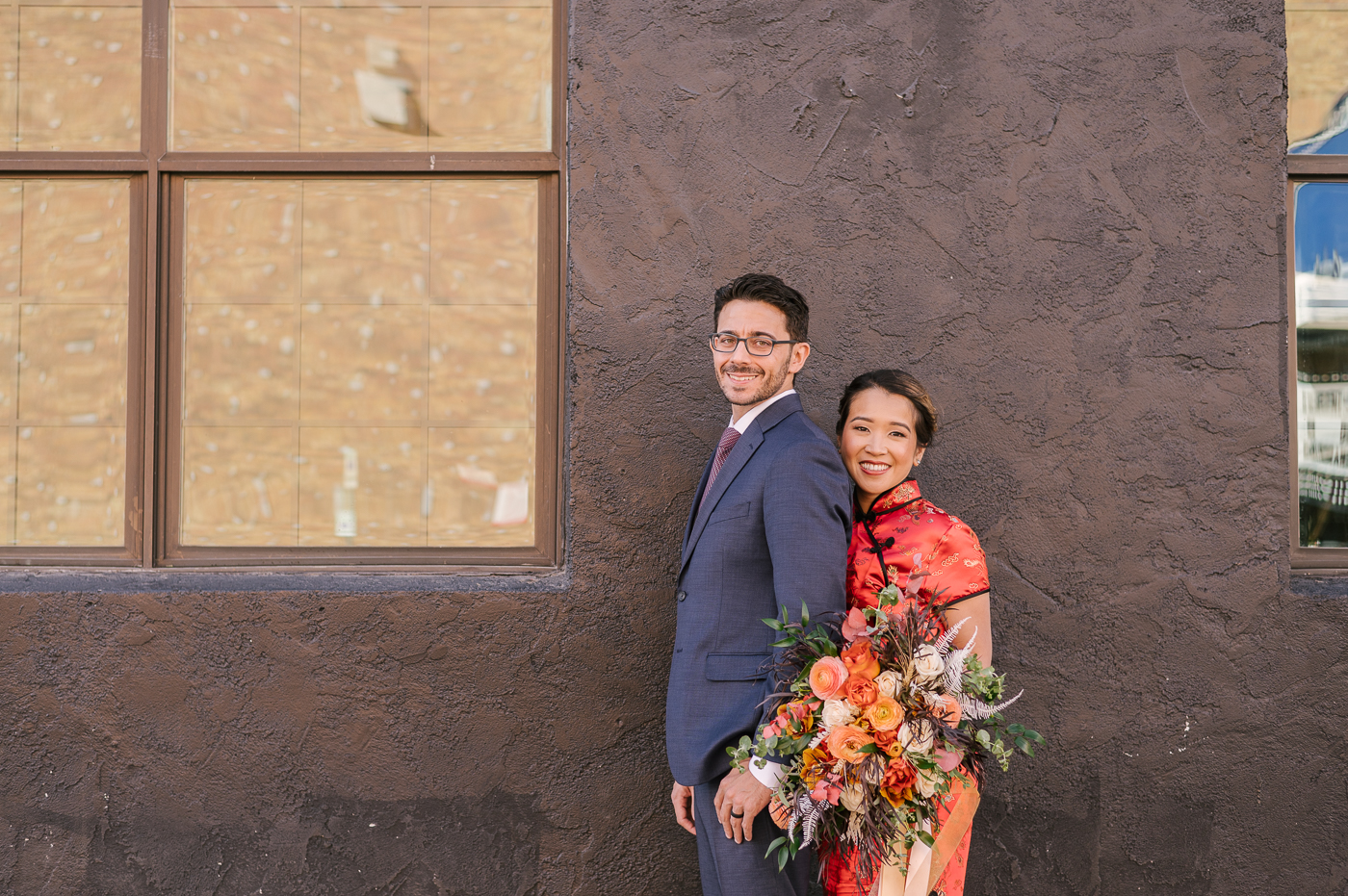 Two Bridal Outfits are Better than One! Newlyweds side by side smiling in front of a black wall that lets their outfits and the wedding bouquet really pop with color 