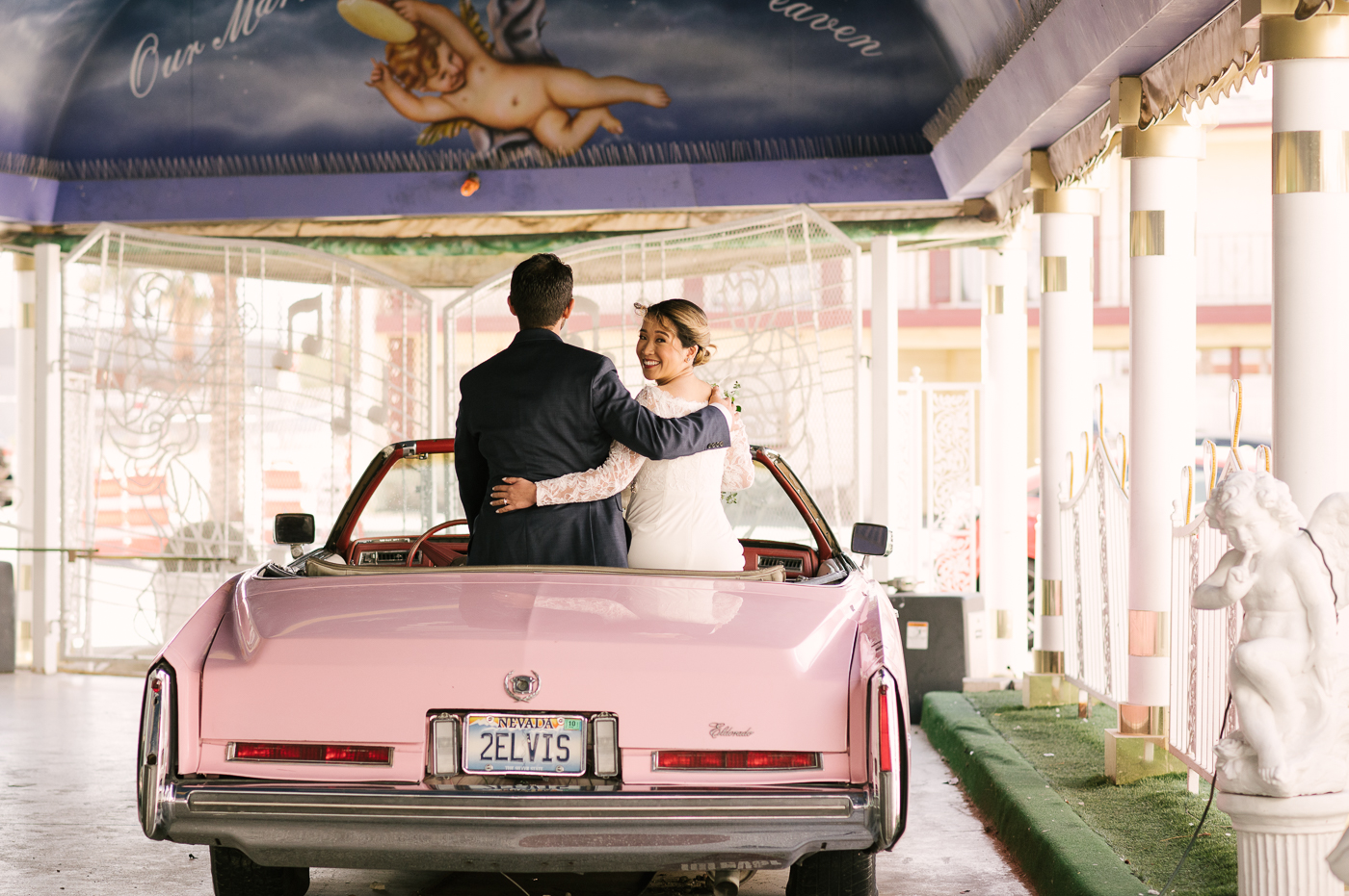 Newlyweds on sitting on the back of the famous Elvis Presley's pink Cadillac with their arms around each other while the bride looks back and smiles.