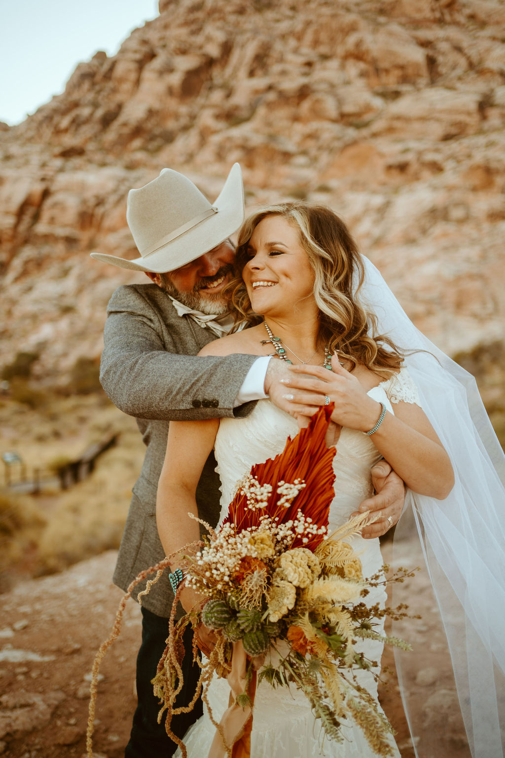 Groom in cowboy hat hugging bride holding dried floral bouquet 