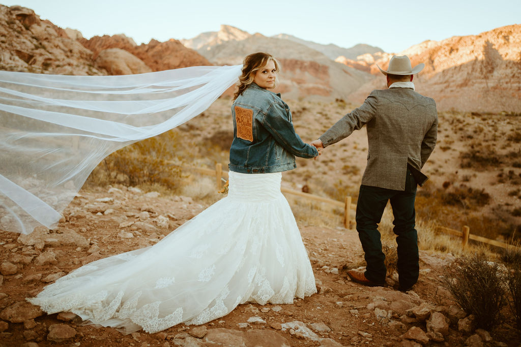 Bride looking back in Custom Jean Jacket and Mermaid Dress with Groom looking off into the distance in Red Rock Canyon 