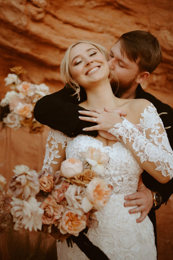  Groom is in an all black suit, bride is wearing an off the shoulders fitted lace wedding dress. With long sleeves and long lace train. Groom holds bride from behind and kissed just under her ear. 