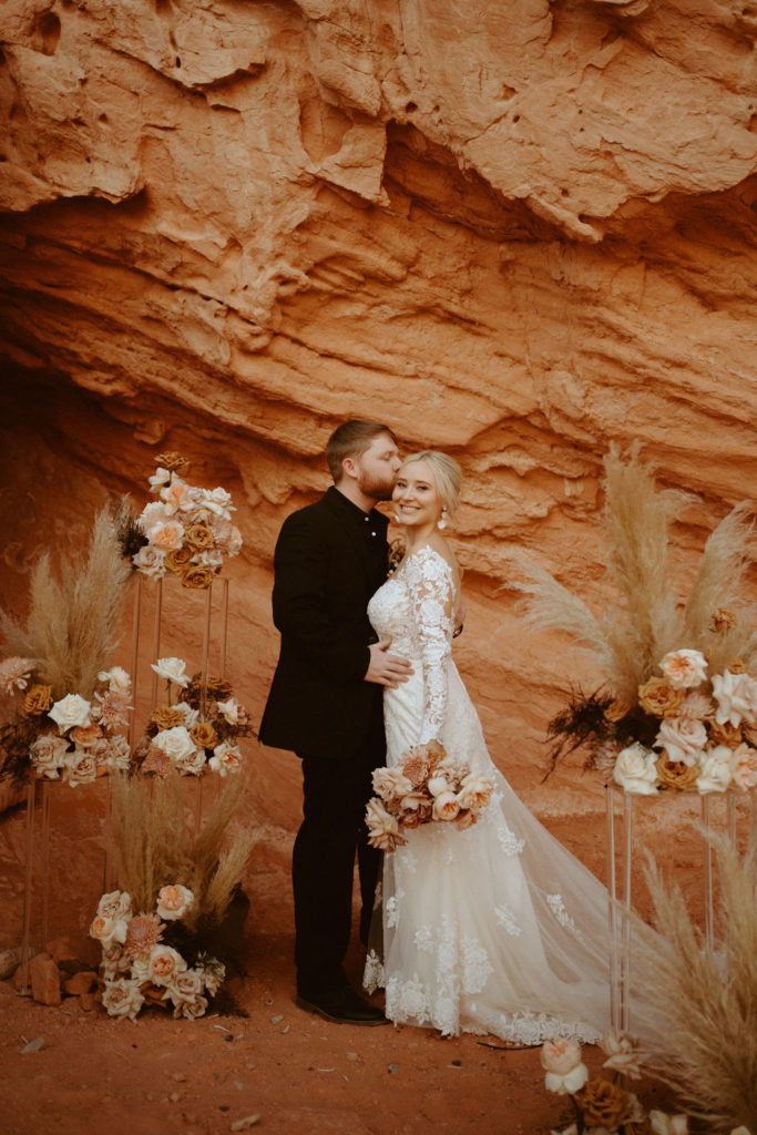  Groom is in an all black suit, bride is wearing an off the shoulders fitted lace wedding dress. With long sleeves and long lace train. Groom kissing his brides temple as she smiles widely at the camera. 