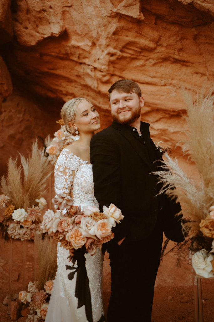 Las Vegas has the Craziest Winds!  Groom is in an all black suit, bride is wearing an off the shoulders fitted lace wedding dress. With long sleeves and long lace train. Bride stands behind the groom holding her bridal bouquet of pink, toffee, and cream.