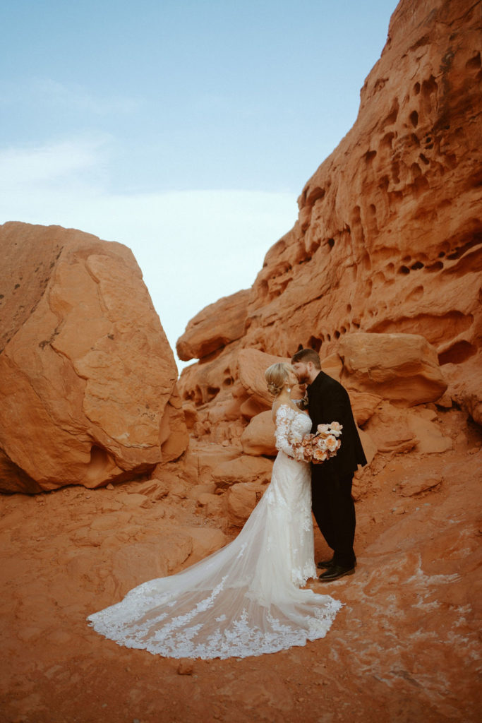 Las Vegas has the Craziest Winds!  Groom is in an all black suit, bride is wearing an off the shoulders fitted lace wedding dress. With long sleeves and long lace train. Sharing a kiss in front of a large red rock boulder. 