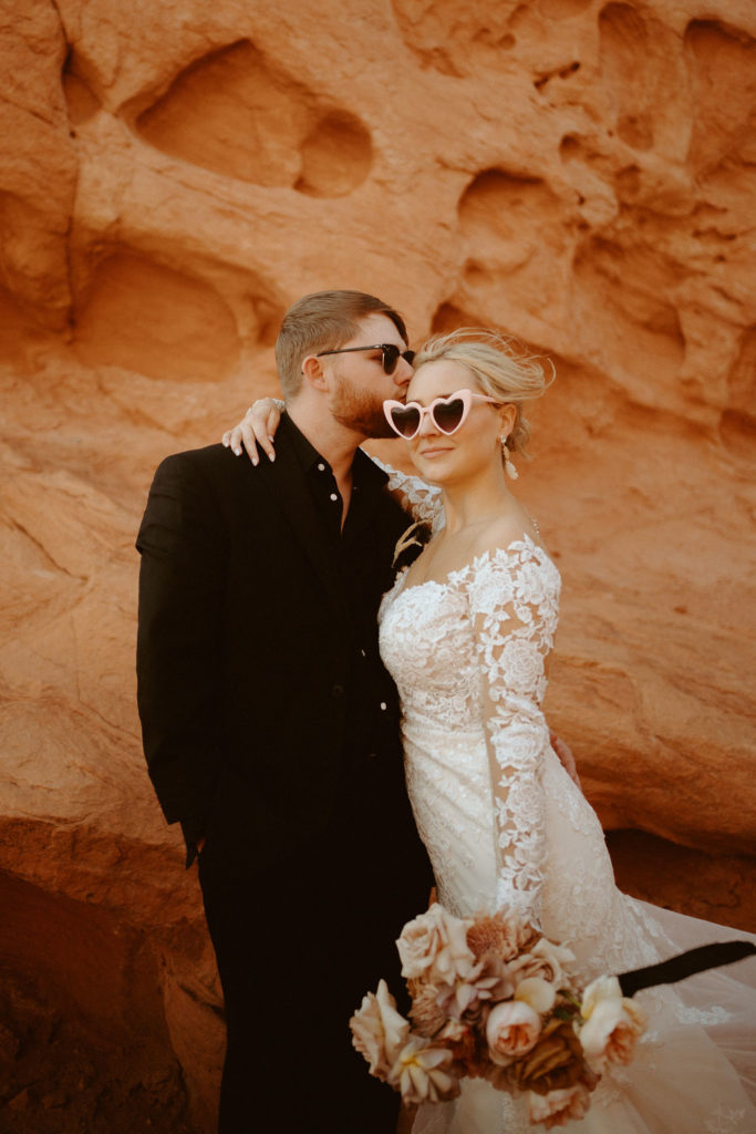  Groom is in an all black suit, bride is wearing an off the shoulders fitted lace wedding dress. With long sleeves and long lace train. Wearing heart sunglasses as the groom kisses the brides temple. 