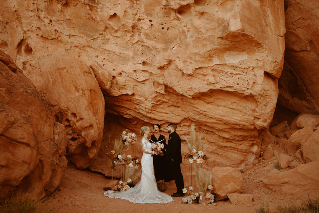 Las Vegas has the Craziest Winds!  Groom is in an all black suit, bride is wearing an off the shoulders fitted lace wedding dress. With long sleeves and long lace train. They stand holding hands for their elopement in the red rocks with ground floral arches surrounding them. 