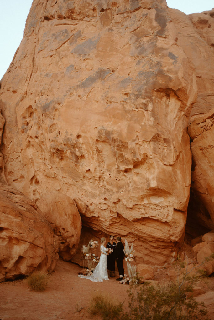  Groom is in an all black suit, bride is wearing an off the shoulders fitted lace wedding dress. With long sleeves and long lace train. Wide dramatic shot of the bride and groom standing at the massive red rocks for their elopement. 