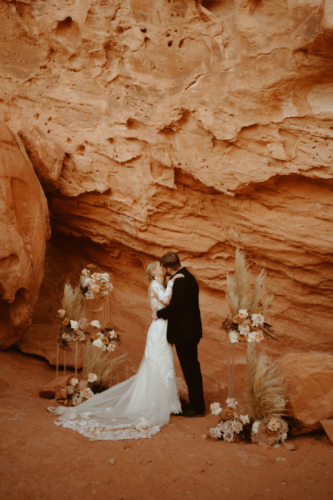 Las Vegas has the Craziest Winds!  Groom is in an all black suit, bride is wearing an off the shoulders fitted lace wedding dress. With long sleeves and long lace train. Having their first kiss as husband and wife. Ground floral arches an either side of the couple. 