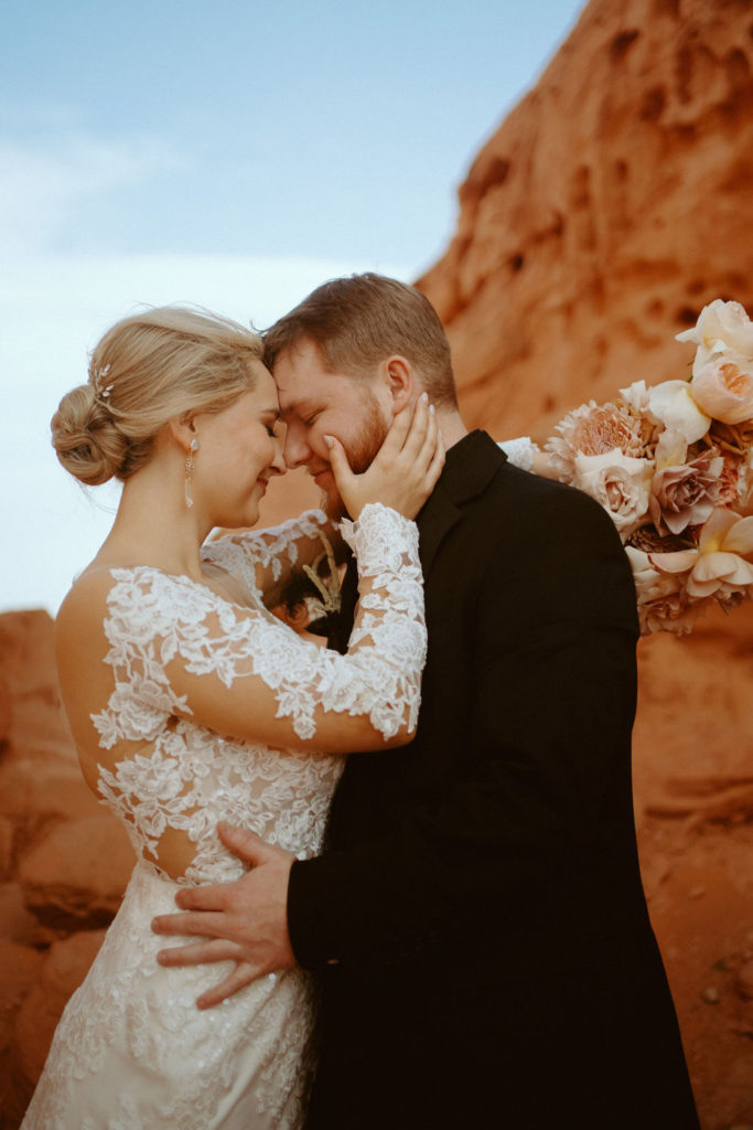  Groom is in an all black suit, bride is wearing an off the shoulders fitted lace wedding dress. With long sleeves and long lace train. Bride holds the grooms face as they press their foreheads together. 
