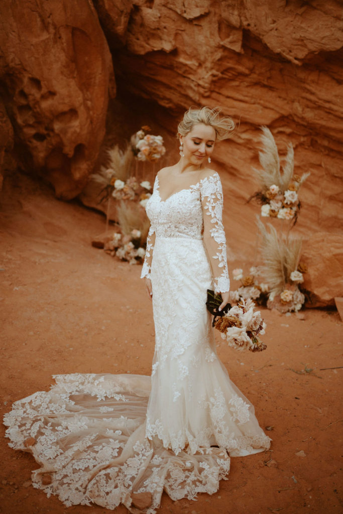 Las Vegas has the Craziest Winds! Bride is wearing an off the shoulders fitted lace wedding dress. With long sleeves and long lace train. Holding onto her bridal bouquet and looking down at the ground, the brides stands just in front of her ground floral arches. 