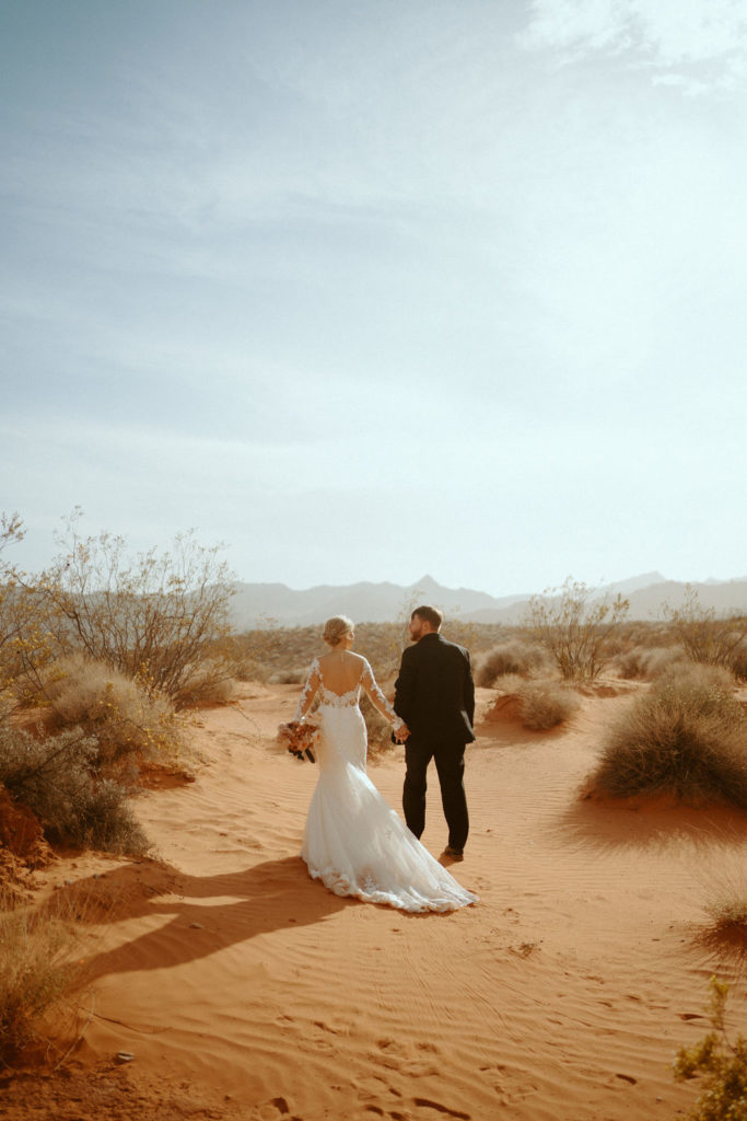 Las Vegas has the Craziest Winds!  Groom is in an all black suit, bride is wearing an off the shoulders fitted lace wedding dress. With long sleeves and long lace train. Walking hand in hand in the Las Vegas desert sand. 