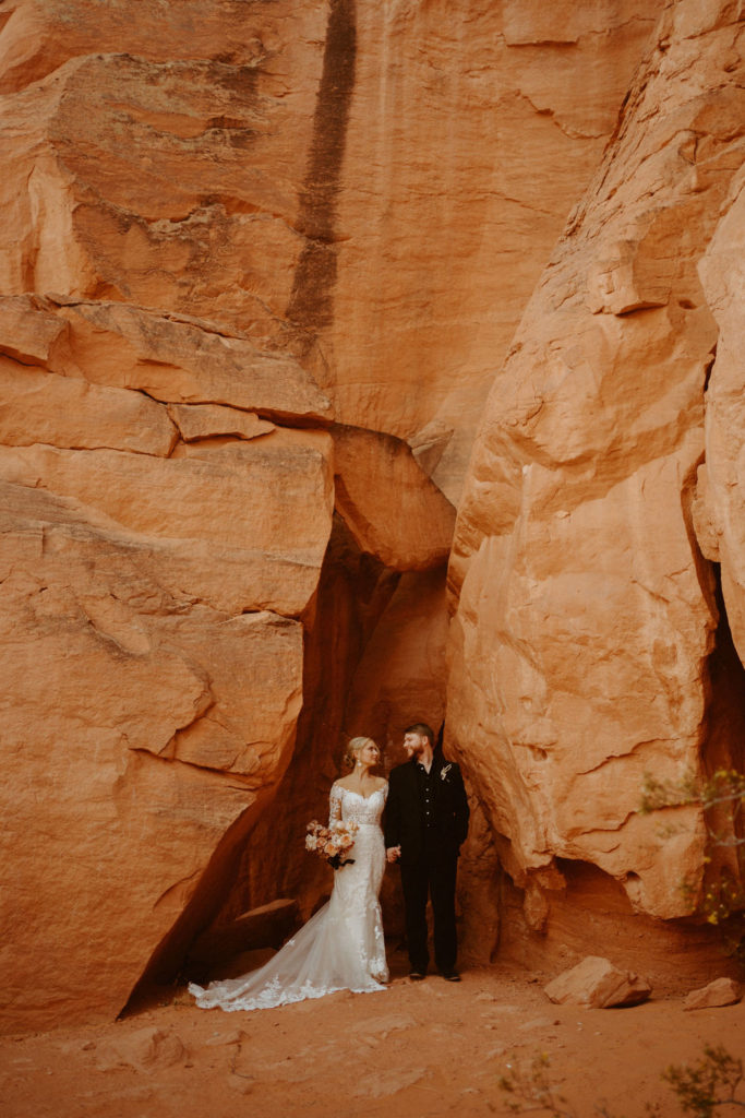Las Vegas has the Craziest Winds!  Groom is in an all black suit, bride is wearing an off the shoulders fitted lace wedding dress. With long sleeves and long lace train. Standing hand in hand for a dramatic wide shot as the couple stands in-between two red rock boulders. 