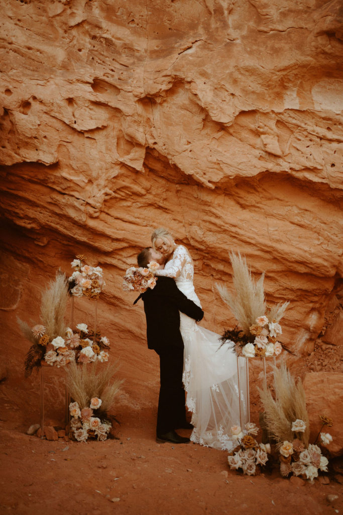  Groom is in an all black suit, bride is wearing an off the shoulders fitted lace wedding dress. With long sleeves and long lace train. Groom holds bride underneath her bottom kissing her cheek as her arms are wrapped around his neck. 