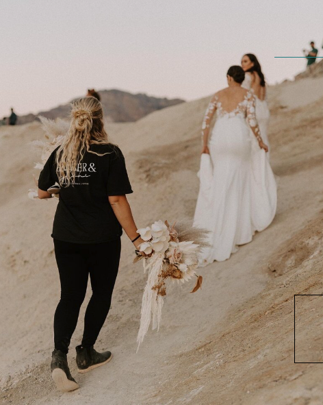 Wedding planner holding bouquet for couple as they walk up desert rocks to the next location for photos, same sex wedding couple, wedding dresses