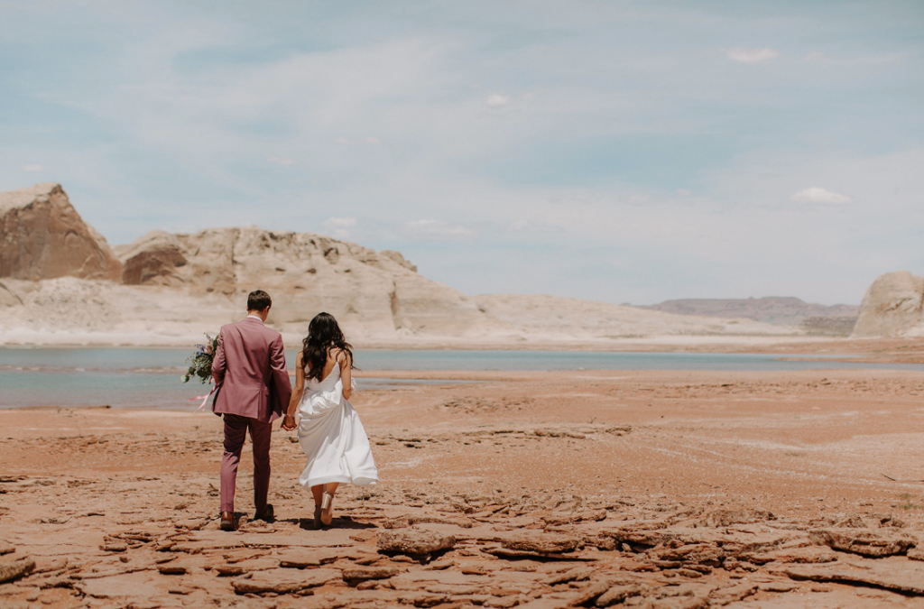 Couple walking from ski boat to the water front of the dried up lake powell. Groom in mauve suit or wedding tux. Bride walking in wedding dress. 
