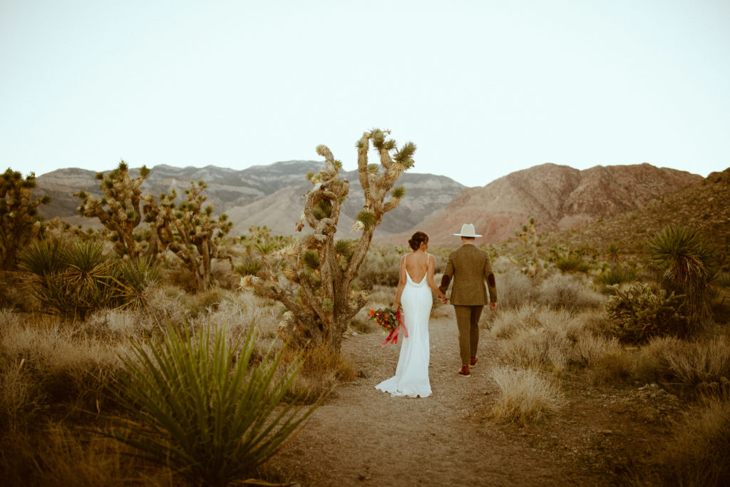 What is a Boho Styled Wedding. The bride in a long white satin dress with a low back holding her colorful bouquet at her side while holding the grooms hand in a tweed suit wearing a cream brim boho style hat both walking out in the middle of the desert at cactus joes.