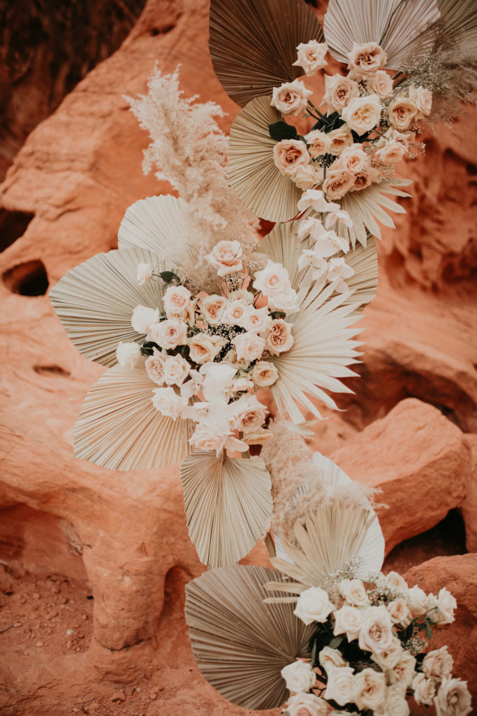 What is a boho styled wedding? Modern boho style floral arrangement with palm leaves rising up a circle arch with cream, white, rose, and toffee colored roses in the middle of valley of fire. 