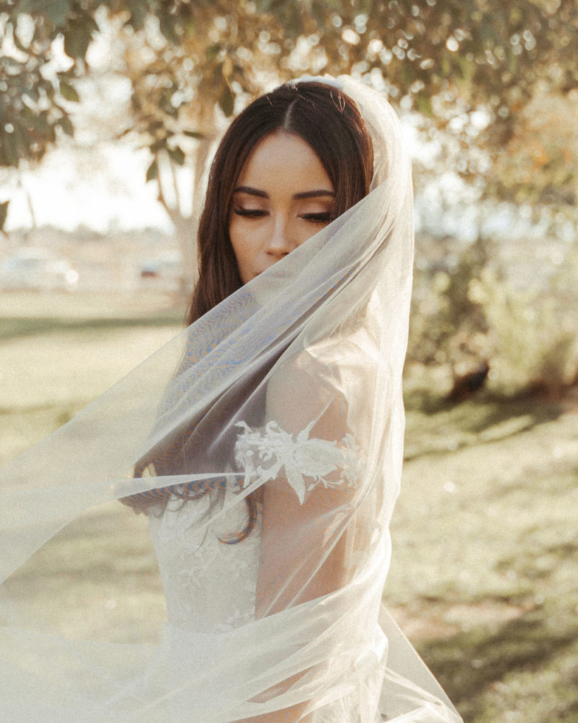 Modern and whimsical bride with an off the shoulder wedding gown with lace embellishments and dramatic beading and a veil blowing in the wind. 
