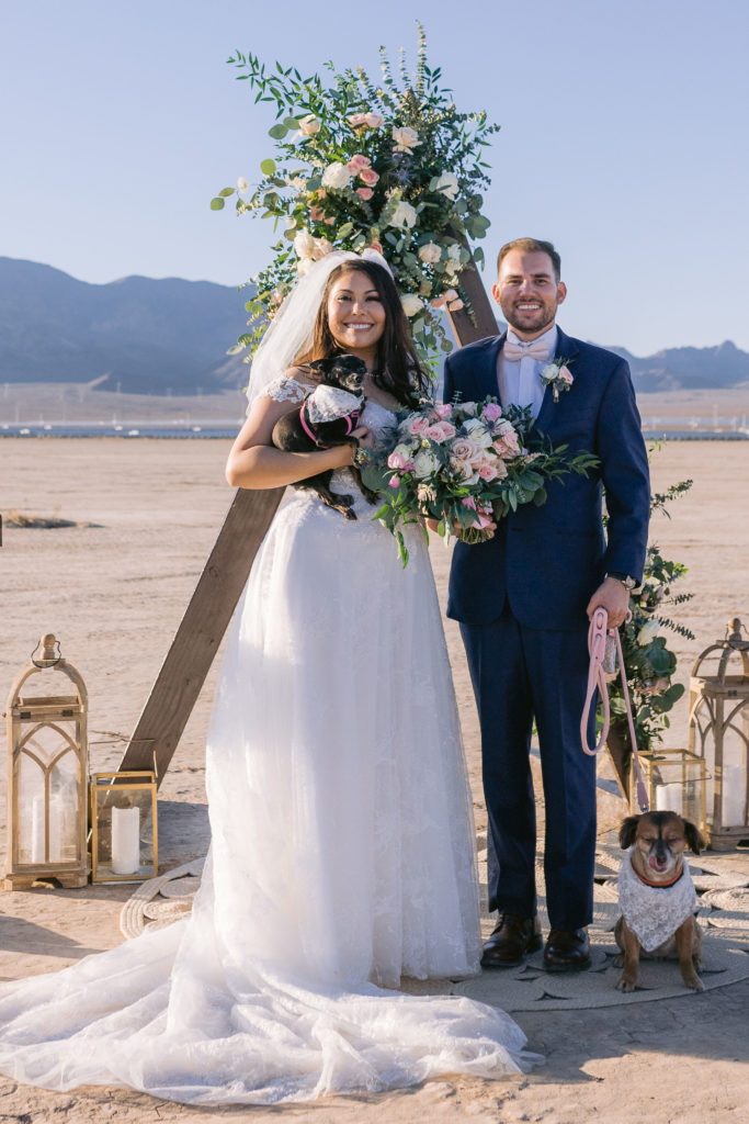 How To Include Your Dog on Your Wedding Day! Newlywed standing in front of a triangle arch draped with flowers on a boho style rug at the altar with both of their dogs. 