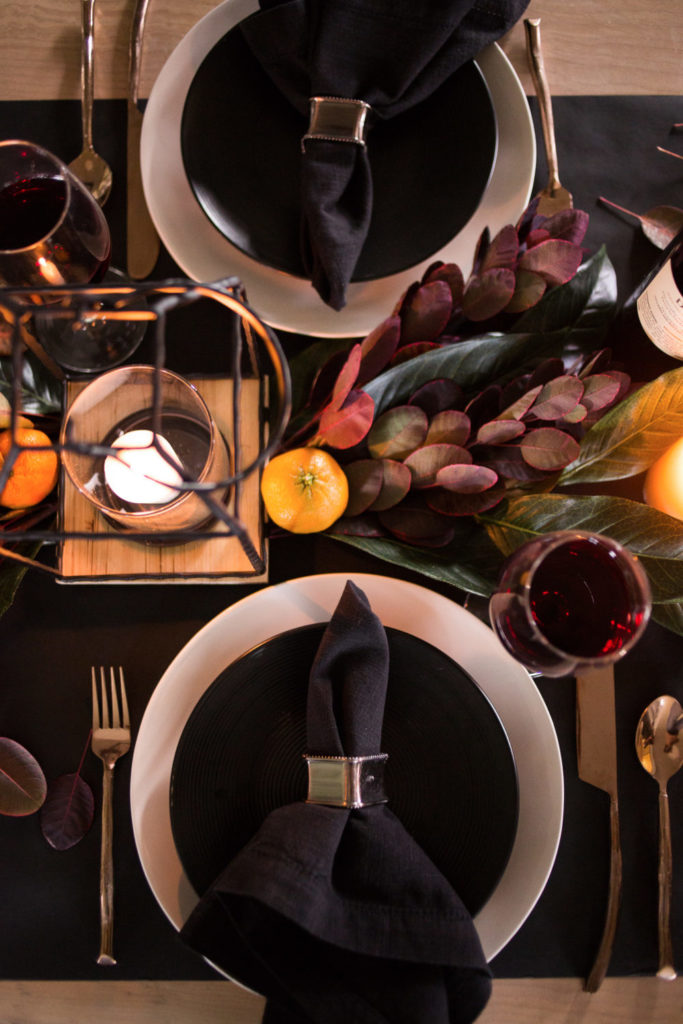How to style the perfect thanksgiving table! Modern and moody thanksgiving table scape. Bright colored fruit mixed in with magnolias and purple foliage and lit candles paired with a white dinner plate, black salad plate and a black folded napkin with a gold napkin ring in the middle. 