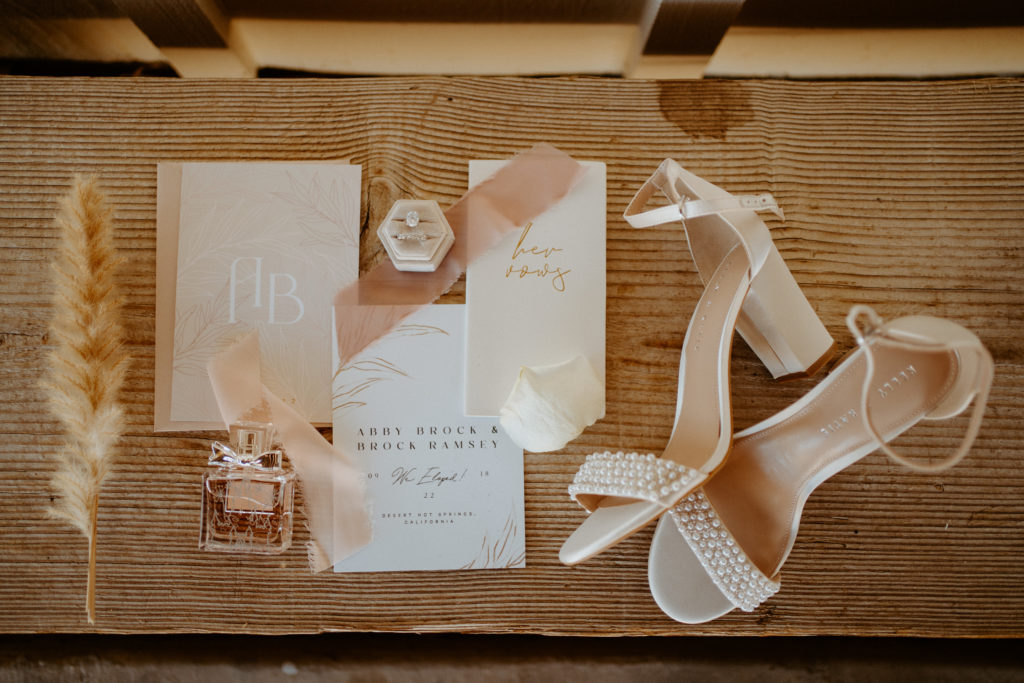 What is a Timeless & Romantic Styled Wedding. Invitation suite flat lay with the brides engagement ring and band, the day of perfume, and nude heels with pearl embellishments. 