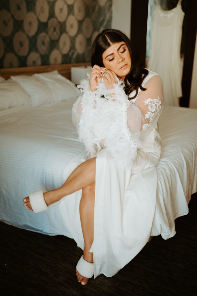 What is a Timeless & Romantic Styled Wedding. Bride getting ready in a silk and lace long robe with feather detailing on the ends of the sleeves and fuzzy white slippers. 