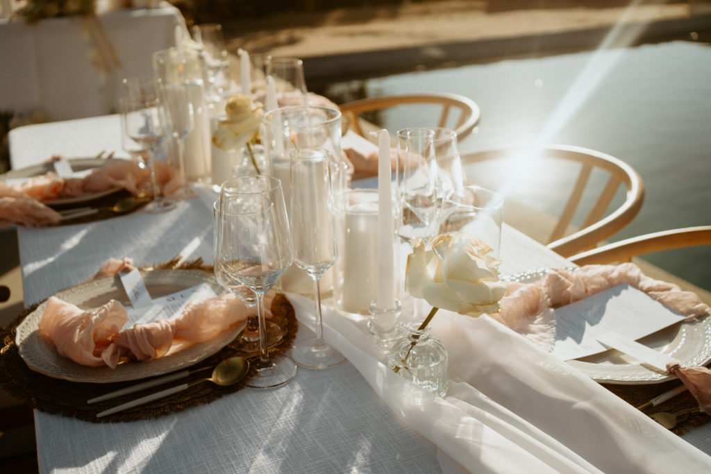 What is a Timeless & Romantic Styled Wedding. Romantic wedding table scape with candles and bud vases with white roses on a long white chiffon table runner. Boho style place mats and cream plating with a blush knot napkin. 