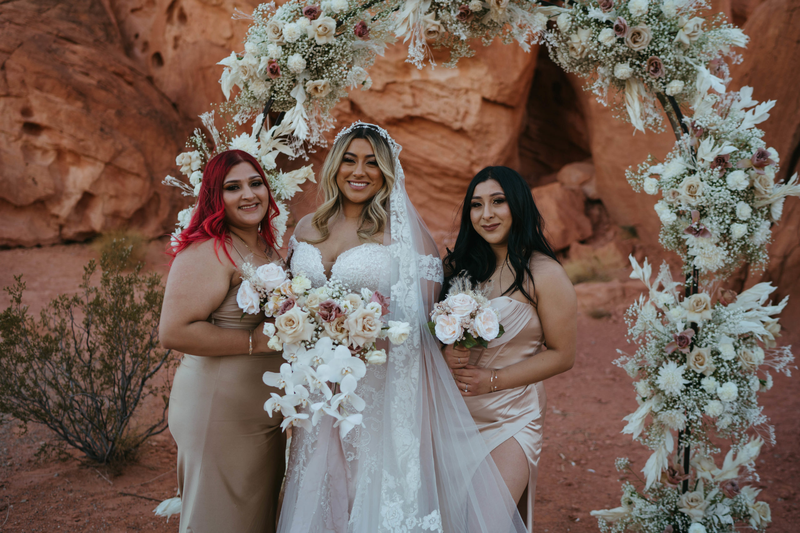 The bride posing with two of her bridesmaids in front of the floral arch in the desert